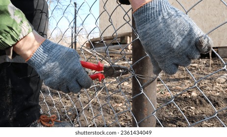 hands in textile gloves with pliers attach galvanized mesh to an iron post using wire in the process of fencing an area on a farm or ranch, securing the metal mesh when installing a barrier