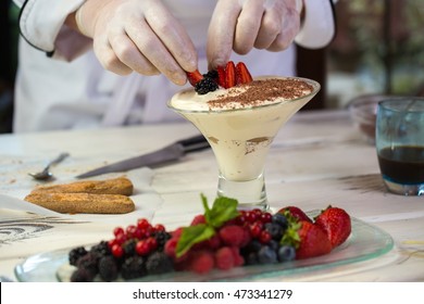 Hands touch pieces of berries. Biscuits and glass with dessert. Fresh strawberries for tiramisu. Chef is decorating sweet dish.