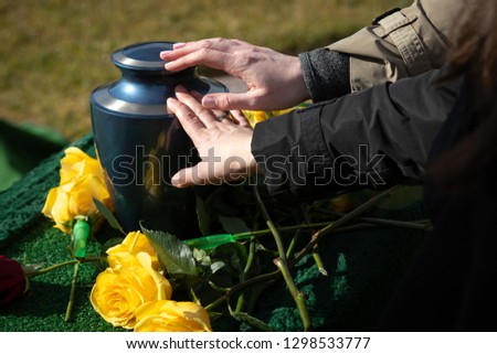 Hands touching a burial urn in a bright outdoor funeral scene, with space for text on the right