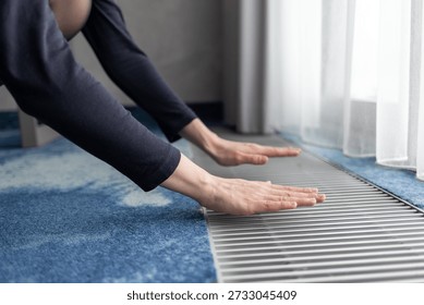 Hands touching a floor convector radiator near a window. Underfloor heating system providing warm air circulation, thermal comfort, and energy efficient heating in a modern interior.

Home heating concept showing heat distribution, indoor comfort, energy saving technology, and efficient space heating during cold seasons.