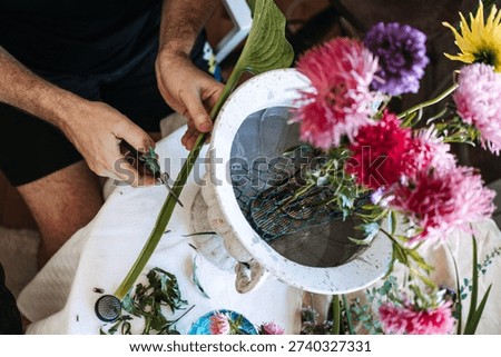Hands trim long green stem with clippers next to white stone urn containing pink asters. nostalgic floral scents, memory triggering flowers, vintage scented garden, comfort flower arranging