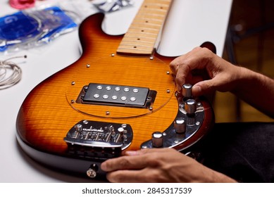 Hands tuning control knobs on sunburst electric guitar laid on workbench, showing pickup, bridge, and chrome hardware with warm lighting and tools blurred in background