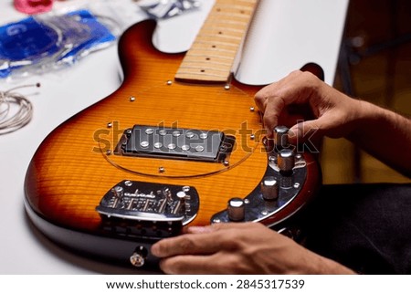 Hands tuning control knobs on sunburst electric guitar laid on workbench, showing pickup, bridge, and chrome hardware with warm lighting and tools blurred in background