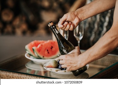 Hands Twist The Muse On A Bottle Of Sparkling Wine. Back On The Glass Table A Plate With Watermelon And Fruit
