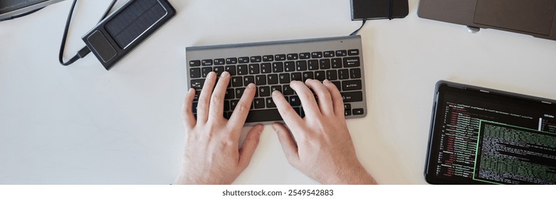 Hands typing on a wireless keyboard surrounded by various gadgets including a smartphone, power bank, and laptop suggesting tech-focused workspace - Powered by Shutterstock