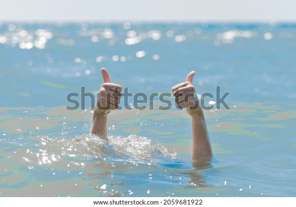 Hands from under the water of a drowning girl shows\
class with a thumb up. Help and urgent rescue of a person during a\
dangerous swimming, sos.