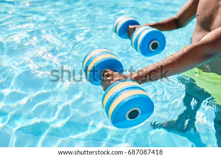 Hands of an unknown black guy holding dumbbells for aqua aerobics standing in the swimming pool on warm summer day