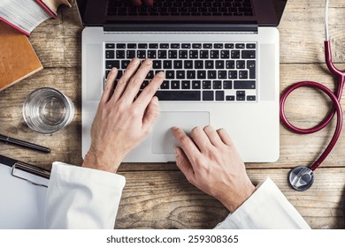 Hands Of Unrecognizable Doctor Writing On A Computer. Wooden Desk Background.