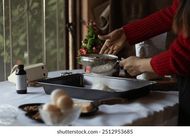 Hands of an unrecognizable woman sifting flour onto a baking tray, surrounded by Christmas ingredients and decorations, while she is preparing some traditional Christmas treats. - Powered by Shutterstock