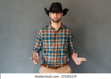 Hands up! Handsome young man wearing cowboy hat and gesturing while standing against grey background 