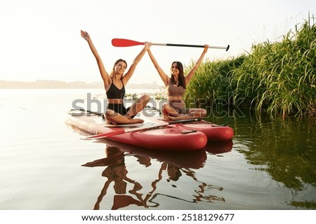 Hands up, smiling and looking forward. Women friends are on sup board on the lake together.