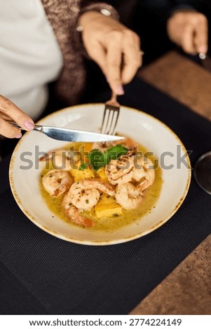 Hands use a fork and knife to cut into a garlic butter shrimp dish served with potatoes at a cozy restaurant table, evoking shared dining, comfort food and relaxed social moments.
