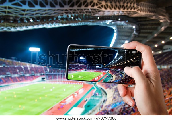 ハンズは サッカー競技場でのスマートフォンのレコード試合を インターネット上のライブストリーミングビデオでフレンドとのソーシャルメディアのコメントチャットに使う の写真素材 今すぐ編集