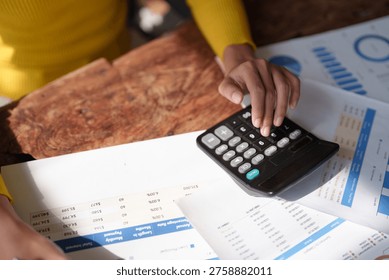 Hands using a calculator to process financial data and accounting reports on a wooden desk, surrounded by paperwork, spreadsheets and charts for budgeting and analysis