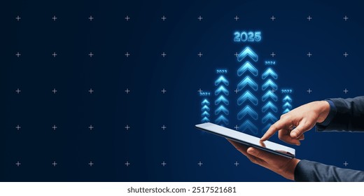 Hands using a digital tablet with glowing arrows pointing up, signifying business growth and goals for 2025 on a dark blue background. Concept of future targets and achievements