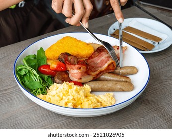 Hands using Knife and fork  Cutting ItalianSausages Full English Breakfast with Crispy Bacon, Scrambled Eggs, Hash Brown, Rocket Salad, Cherry Tomatoes, Mushrooms, and Brown and White Sugar Packets