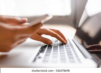 Hands mit Laptop und Telefon für Online-Shopping, Nahaufnahme vor dem Fenster in sanftem Licht