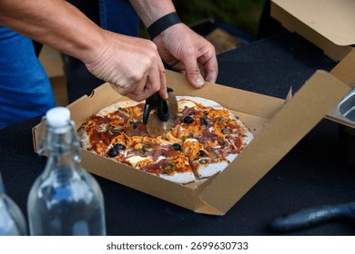 Man’s hands using a pizza cutter to slice a personal size wood fired pizza in a carboard box, cooked fresh outside in a food truck
