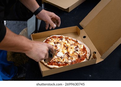Man’s hands using a pizza cutter to slice a personal size wood fired pizza in a carboard box, cooked fresh outside in a food truck

