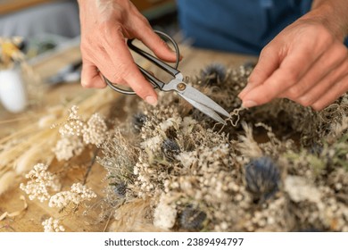 Hands using scissors to trim a dried flower wreath on a wooden workshop table