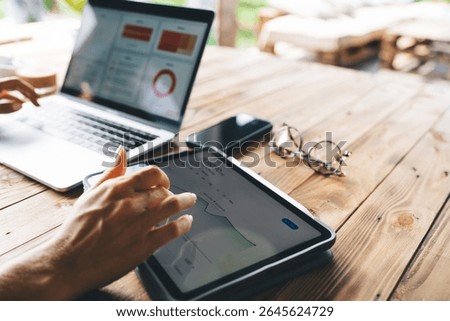 Hands using tablet for real-time financial stats next to open laptop with business charts, glasses and phone on rustic desk, remote work concept in creative natural environment