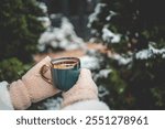 Hands in warm mittens holding a steaming cup of tea in a green mug with a golden rim, surrounded by snow-covered trees in a winter garden setting.