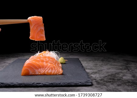 The hands were holding the chopsticks to hold the salmon sashimi, which was arranged on a black stone plate on a old table, with copy space.