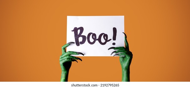 Hands Of Witch Holding Paper With Text BOO On Orange Background. Halloween Celebration