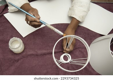 Hands of a woman applying glue with a long brush to the metal of a lampshade on a purple tablecloth