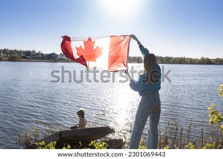 in hands of woman, Canadian flag is fluttering on a sunny day against the sky and a lake, a child is sitting nearby. Travel, holidays, immigration. Canada Day. Pride, freedom, national symbol