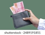 Hands of woman holding black wallet and piles of Indonesian rupiah banknotes for Rp.100,000 isolated on green background. Indonesian rupiah currency