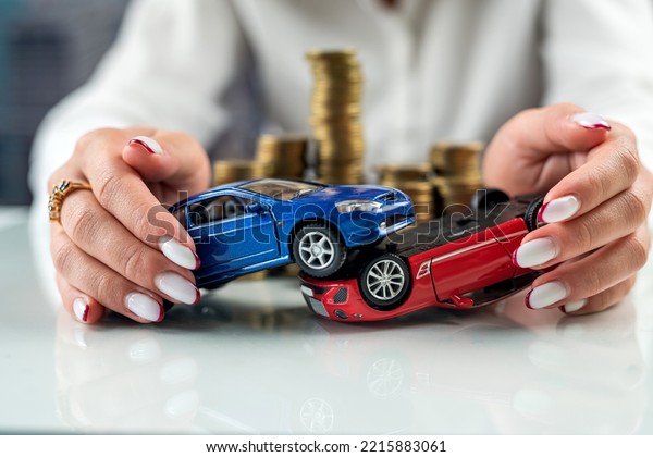 Hands of a woman holding two car models simulating\
an accident near a mountain of coins. Car insurance. Car accident\
driving safety