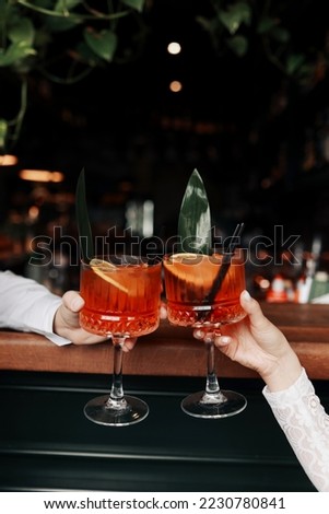 hands of woman and man are clinking, cheers with glasses of Spritz cocktail. Couple celebrating wedding, anniversary with Aperol spritz cocktails, with orange and greens Refreshing alcoholic drink.