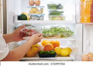 Hands of woman opening fridge door and putting package of fresh ripe strawberries in it