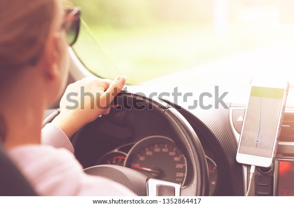 Hands of a\
woman with a steering wheel, Driving a car on a smartphone\
navigator. Photo of a young woman driving a car. Rear view and rear\
view of a young beautiful woman driving a\
car.