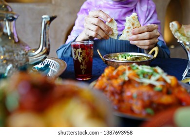 Hands Of Woman In Traditional Muslim Clothing Tearing Naan Bread Before Eating With Palak Paneer (Indian Dish Featuring Spinach And Cheese) On Dining Table With Blurred Aloo Gobi And Moroccan Tea Set.