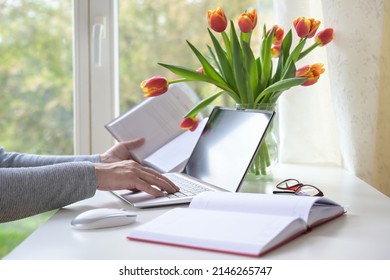 Hands of a woman typing on a laptop computer, books, mouse and tulip bouquet on a white desk with by the window, study or home office business, copy space, selected focus, narrow depth of field