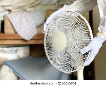 Hands With Work Gloves Hold A Fan In Front Of An Overflowing Closet