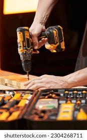 The hands of a worker screw a self-tapping screw into a wooden blank using an electric drill-driver with a replaceable battery. there is a tool box on the table