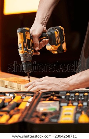 The hands of a worker screw a self-tapping screw into a wooden blank using an electric drill-driver with a replaceable battery. there is a tool box on the table