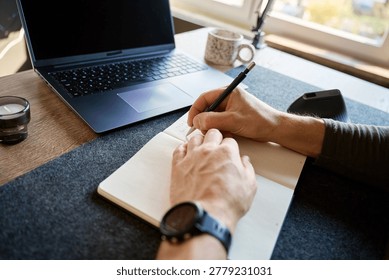 Hands writing notes in notebook beside laptop and smartphone on desk in home office. Concept of planning, remote work, productivity and daily organization