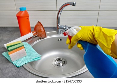 Hands in yellow rubber gloves hold a spray bottle with cleaning agent against the background of detergents and a kitchen sink. Kitchen and sewer cleaning work