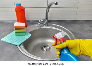 Hands in yellow rubber gloves hold a spray bottle with cleaning agent against the background of cleaning products and a round metal kitchen sink. Kitchen cleaning