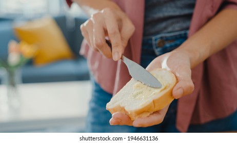 Hands Of Young Asian Woman Chef Spreading Butter On Rustic Rye Bread With Metal Knife On Wooden Board On Kitchen Table In House. Fresh Homemade Bread Production, Healthy Eating And Traditional Bakery.