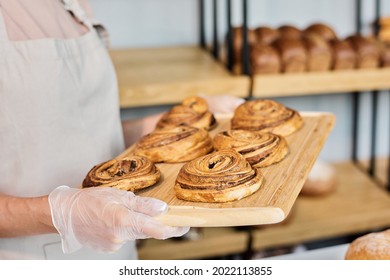 Hands of young baker holding wooden tray with fresh appetizing pastry against display with bread