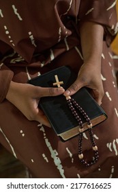 Hands Of Young Black Woman In Brown Dress Holding Bible And Rosary Beads With Small Wooden Cross While Attending Church Service