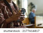 Hands of young black woman with Holy Bible and rosary beads with small wooden cross standing in church and praying after sermon