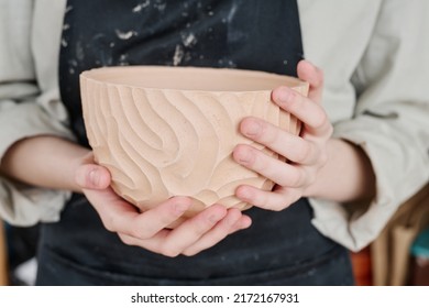 Hands of young creative female artisan or potter in workwear holding handmade clay bowl with carved ornaments on side surface