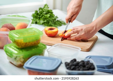 Hands Of Young Female Chopping Fresh Peaches On Wooden Board While Preparing Fruits And Vegetables For Freezing. Plastic Containers With Raw Cut Vegetables For Freezing On Kitchen Table.