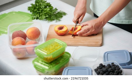 Hands Of Young Female Chopping Fresh Peaches On Wooden Board While Preparing Fruits And Vegetables For Freezing. Stack Of Plastic Containers With Raw Cut Vegetables For Freezing On Kitchen Table.